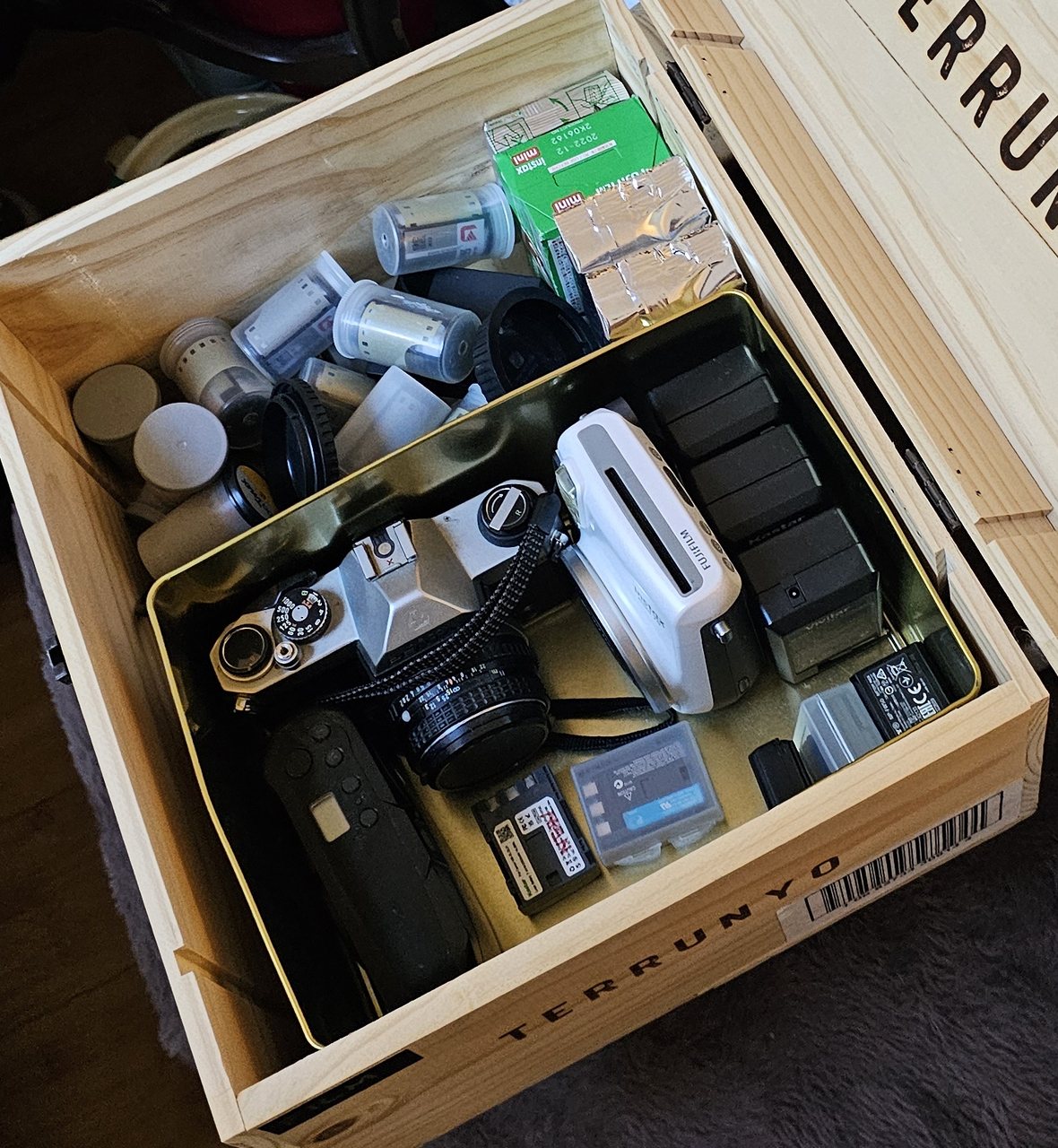 An open wooden crate packed with film cameras, loose rolls of film, an instant camera and spare batteries.